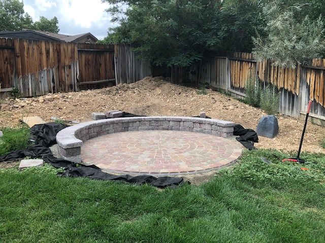 A round paver patio with a seat wall being built in a backyard, liner laid out beside it