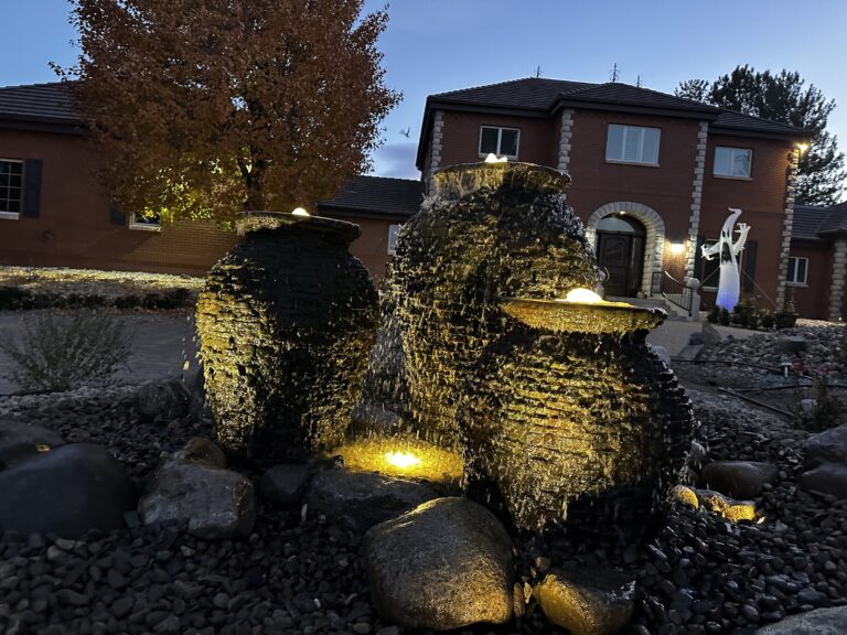 Three stacked-slate urn fountains glowing with landscape lights at dusk in front of a brick house